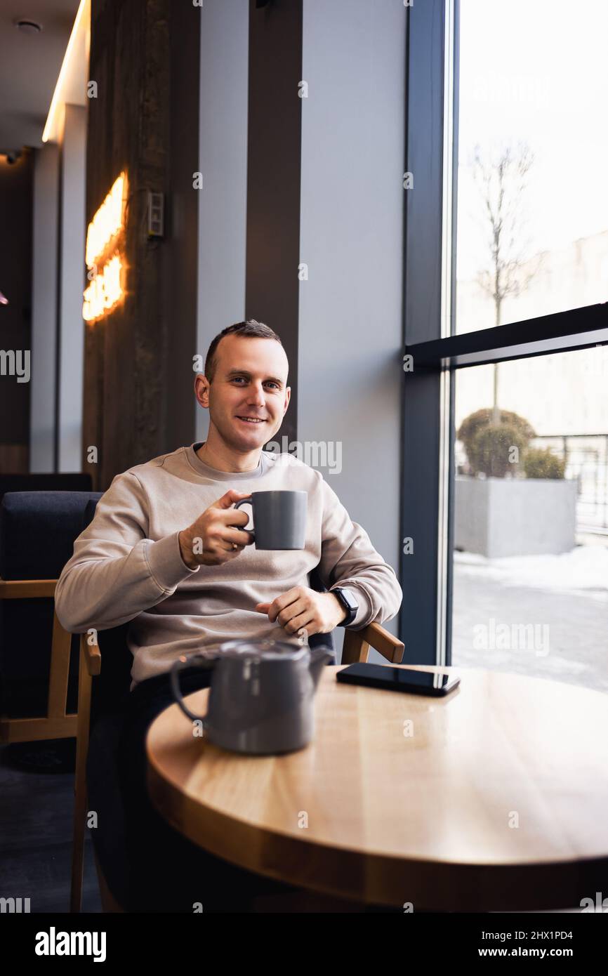 An attractive lonely man sits at a cafe table, he drinks fragrant hot tea. Smiling guy in casual clothes sits in a cafe and drinks coffee Stock Photo