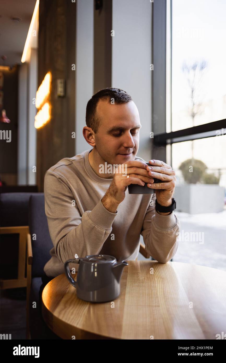 An attractive lonely man sits at a cafe table, he drinks fragrant hot tea. Smiling guy in casual clothes sits in a cafe and drinks coffee Stock Photo