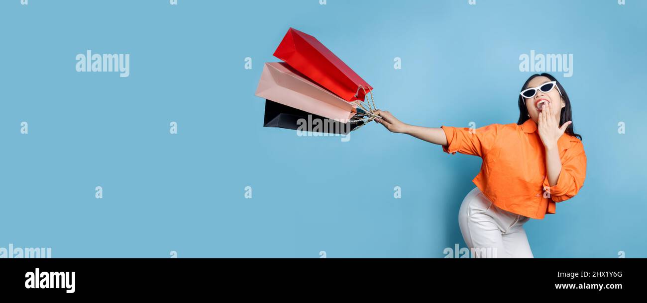 Young fresh and cheerful Asian woman in orange shirt holding and get shopping bag with big smiled on blue background, shopping on holiday and flash se Stock Photo