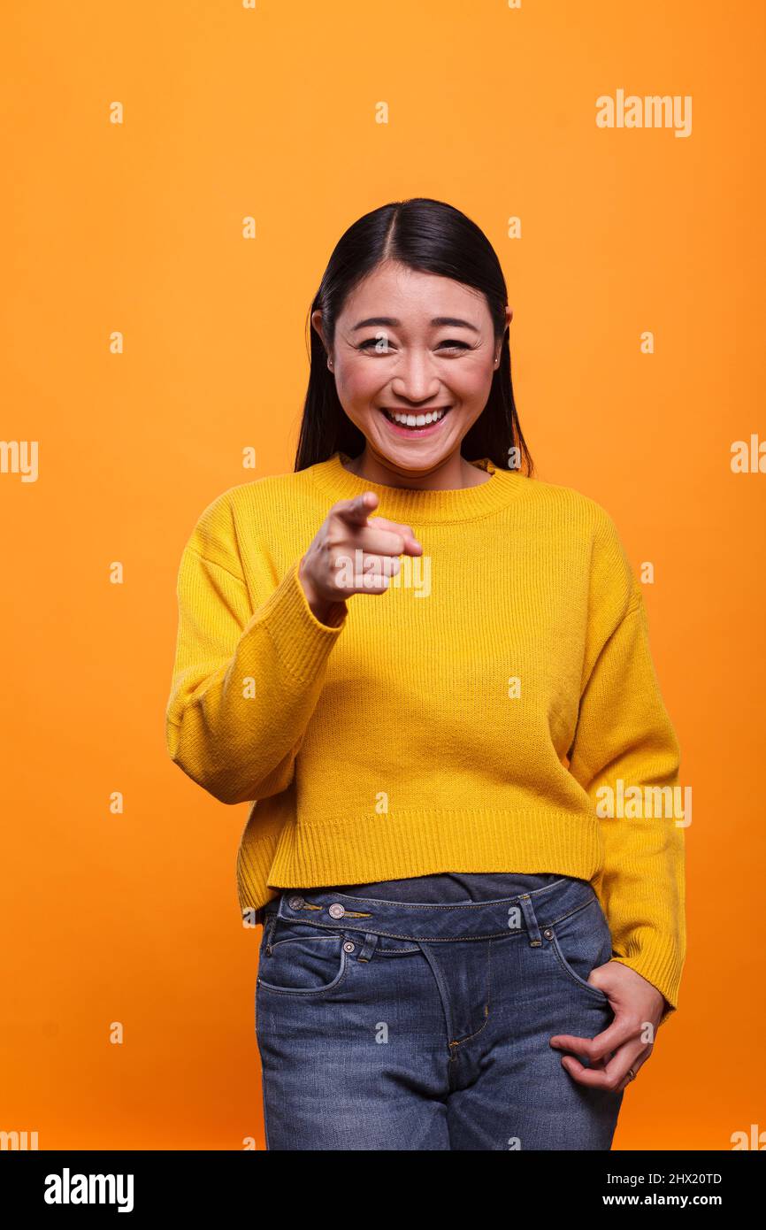 Happy excited beautiful woman wearing yellow sweater pointing index finger at camera on orange background. Joyful positive cute young adult person pointing forefinger to camera. Stock Photo
