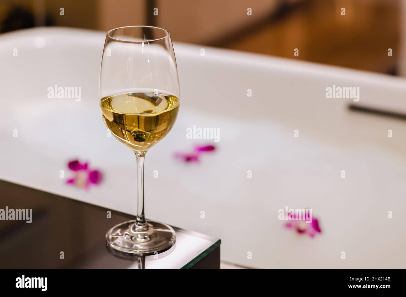 A glass of white wine puts on table beside bathtub that have white bubble and orchid flowers. Relaxing at home concept. Stock Photo