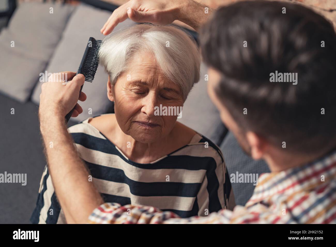 sad grandmother with closed eyes getting her coiffure fixed by her middle-aged millennial son. High quality photo Stock Photo