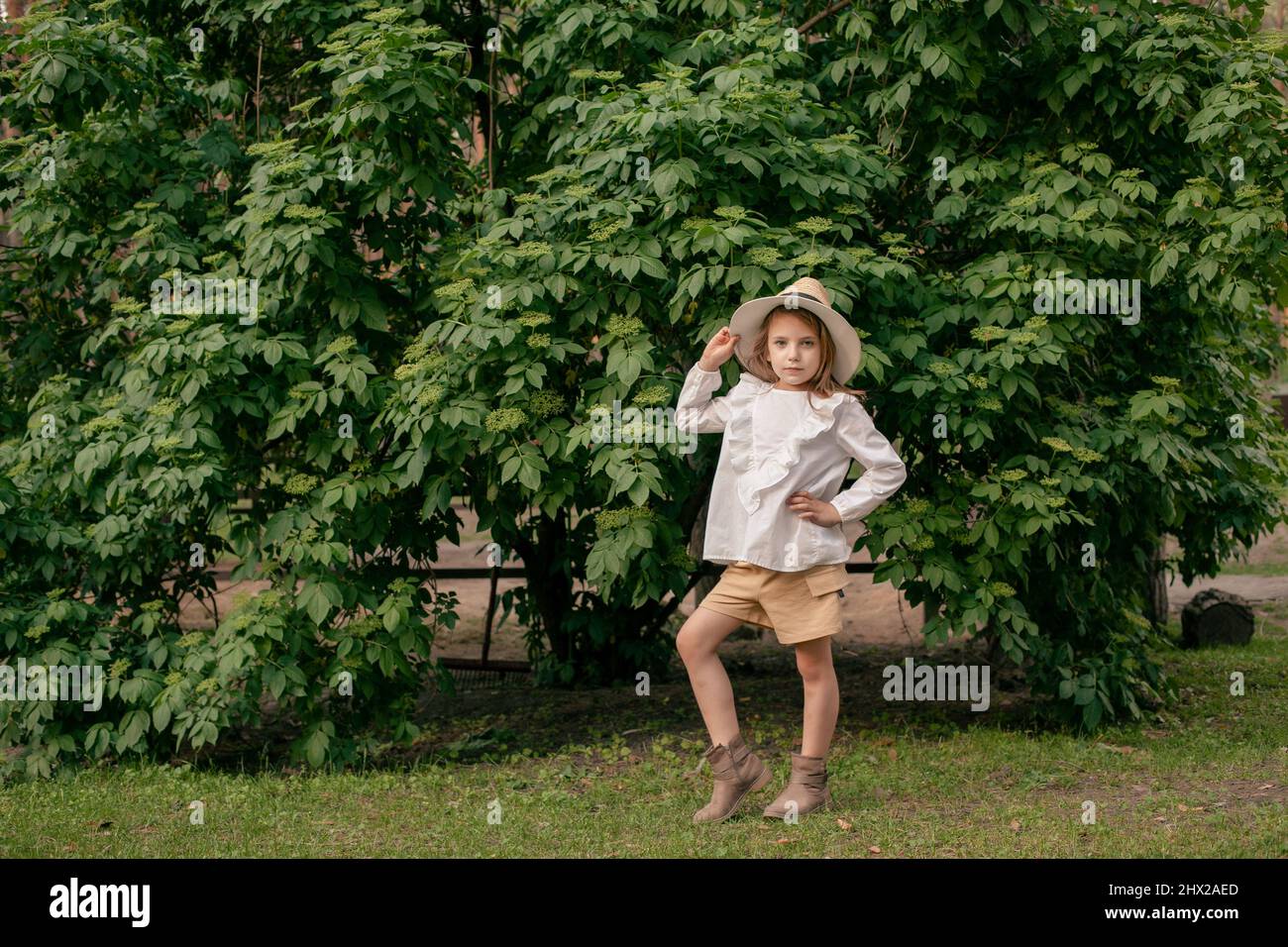Stylish preteen girl standing near green bush in park on summer day ...