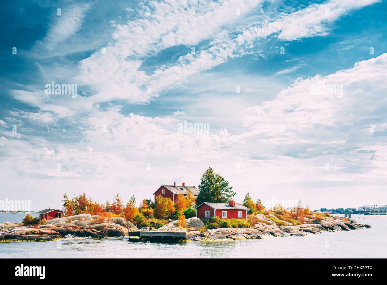 Suomi Or Finland. Beautiful Red Finnish Wooden Log Cabin House On Rocky Island Coast In Summer Sunny Evening. Lake Or River Landscape. Tiny Rocky Stock Photo