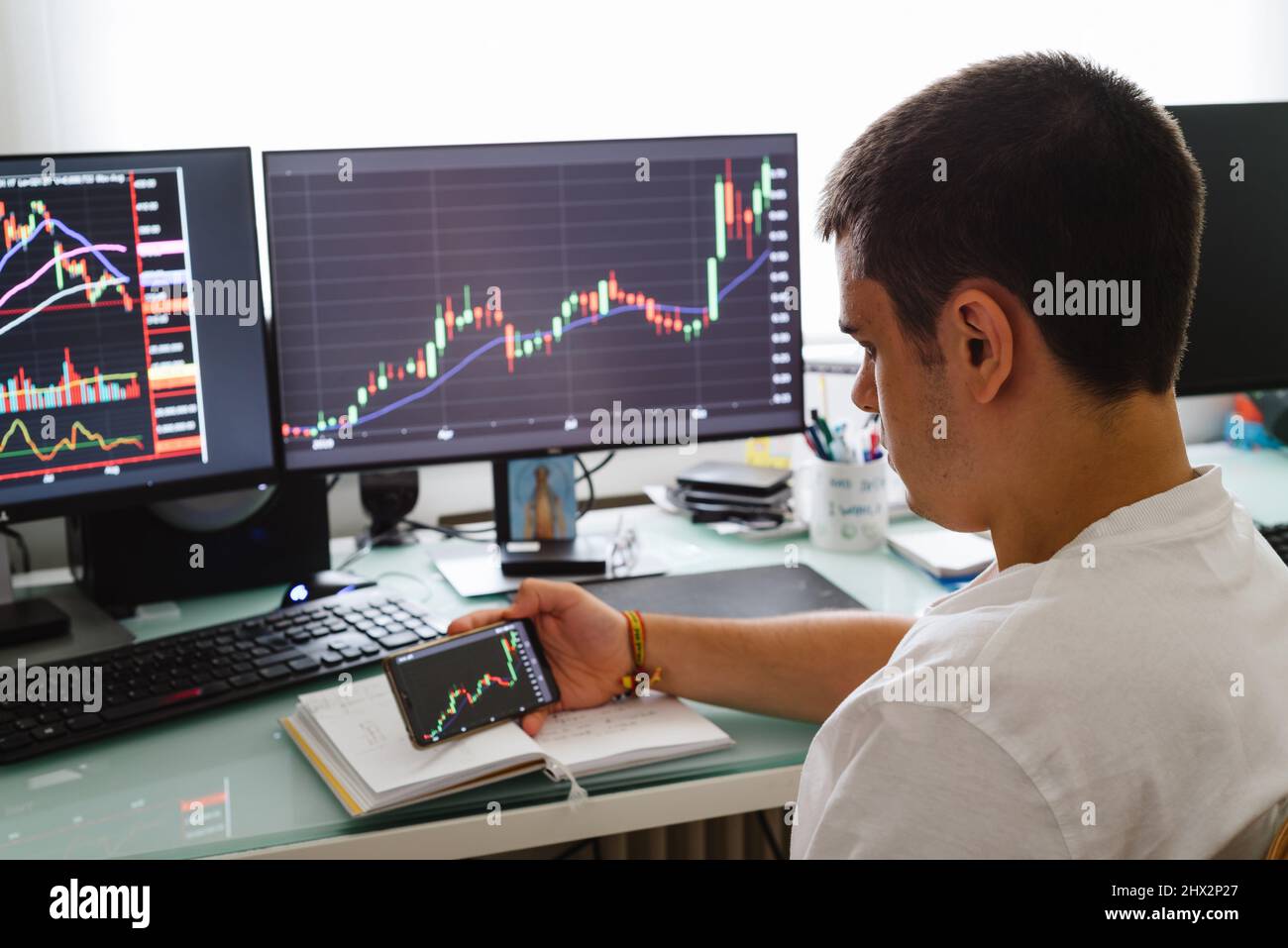 Busy working day at home. Side view of successful young trader in casual wear working with charts and market reports on computer screens in his home. Stock Photo
