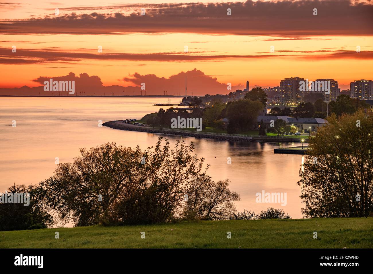 Fiery sunset over a city on a lake. An island dotted with tall wind turbines is visible in background. Stock Photo