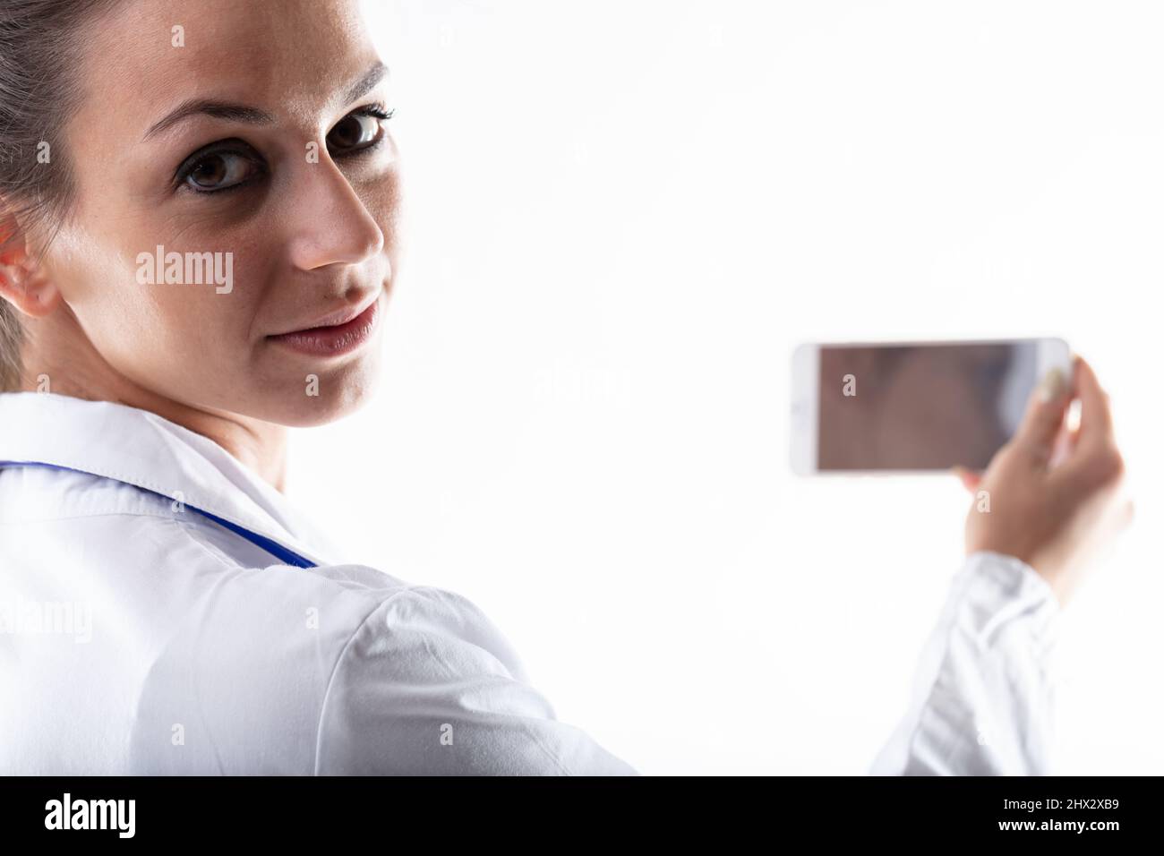 Doctor holding up her mobile phone and looking back at the camera over her shoulder in a cropped portrait with focus to her face Stock Photo