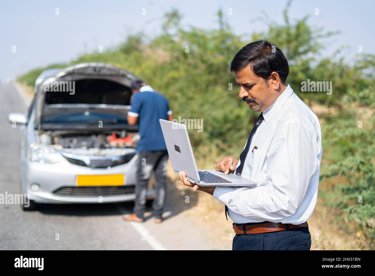 Businessman on roadside busy working on laptop in front of broken car while mechanic buy reparing car - concept of mobile car service, busy work Stock Photo