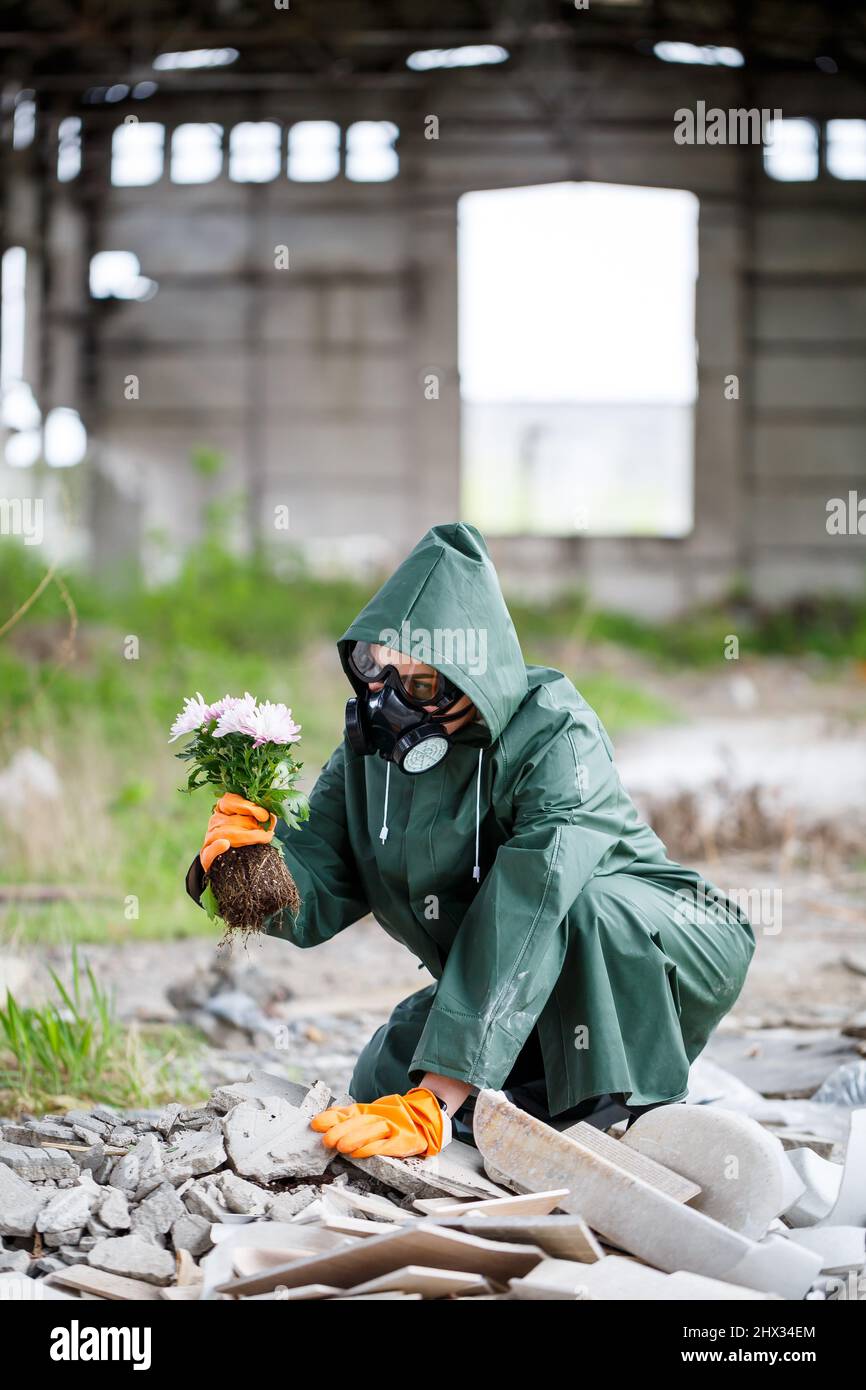 A man in a raincoat and gas mask collects a flower from a scorched, toxic land. Air pollution concept. Ecological catastrophy. Stock Photo