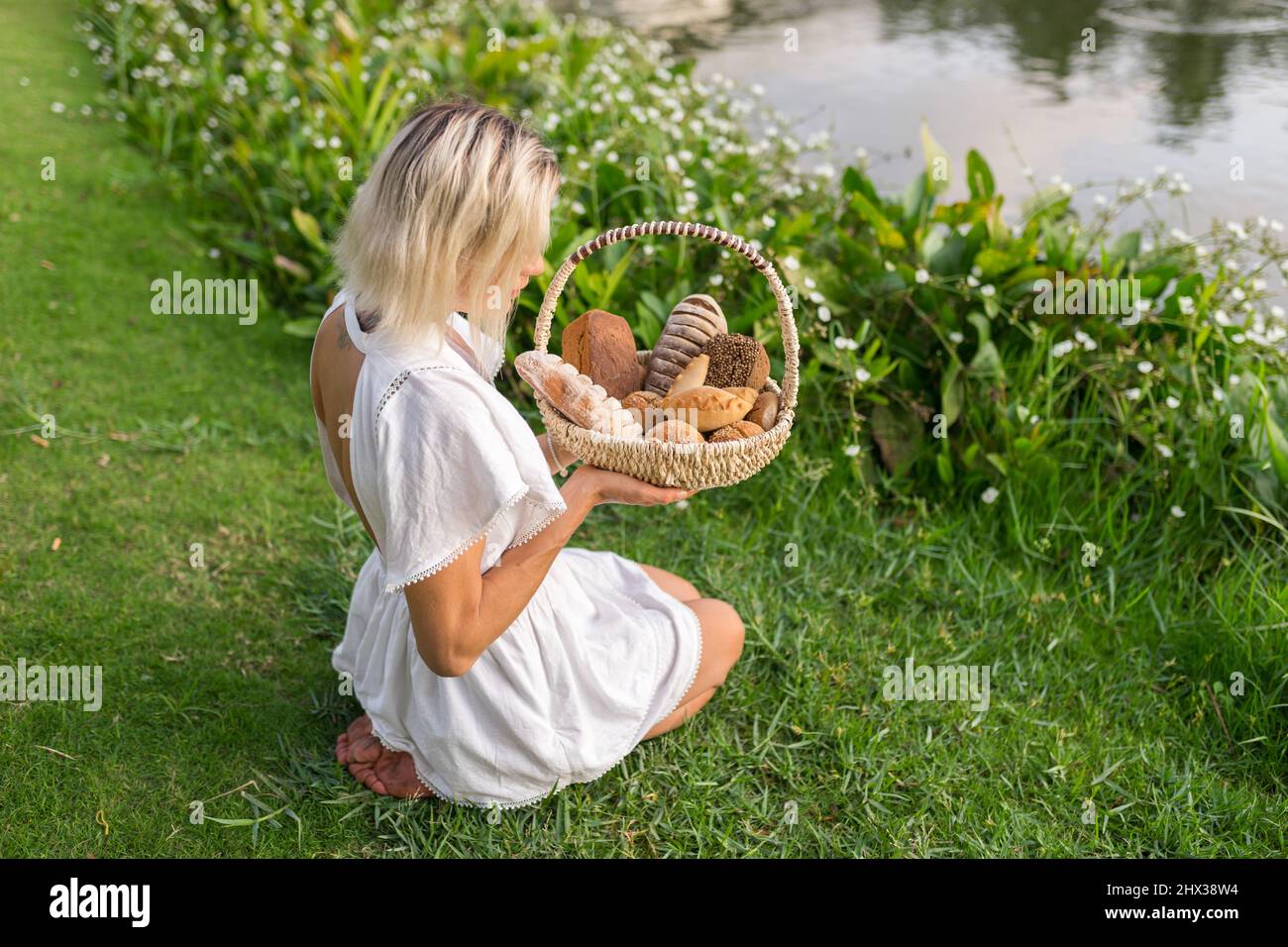 Young woman in white dress sitting on the grass with basket with assorti of brown and white homemade bread. High quality photo Stock Photo