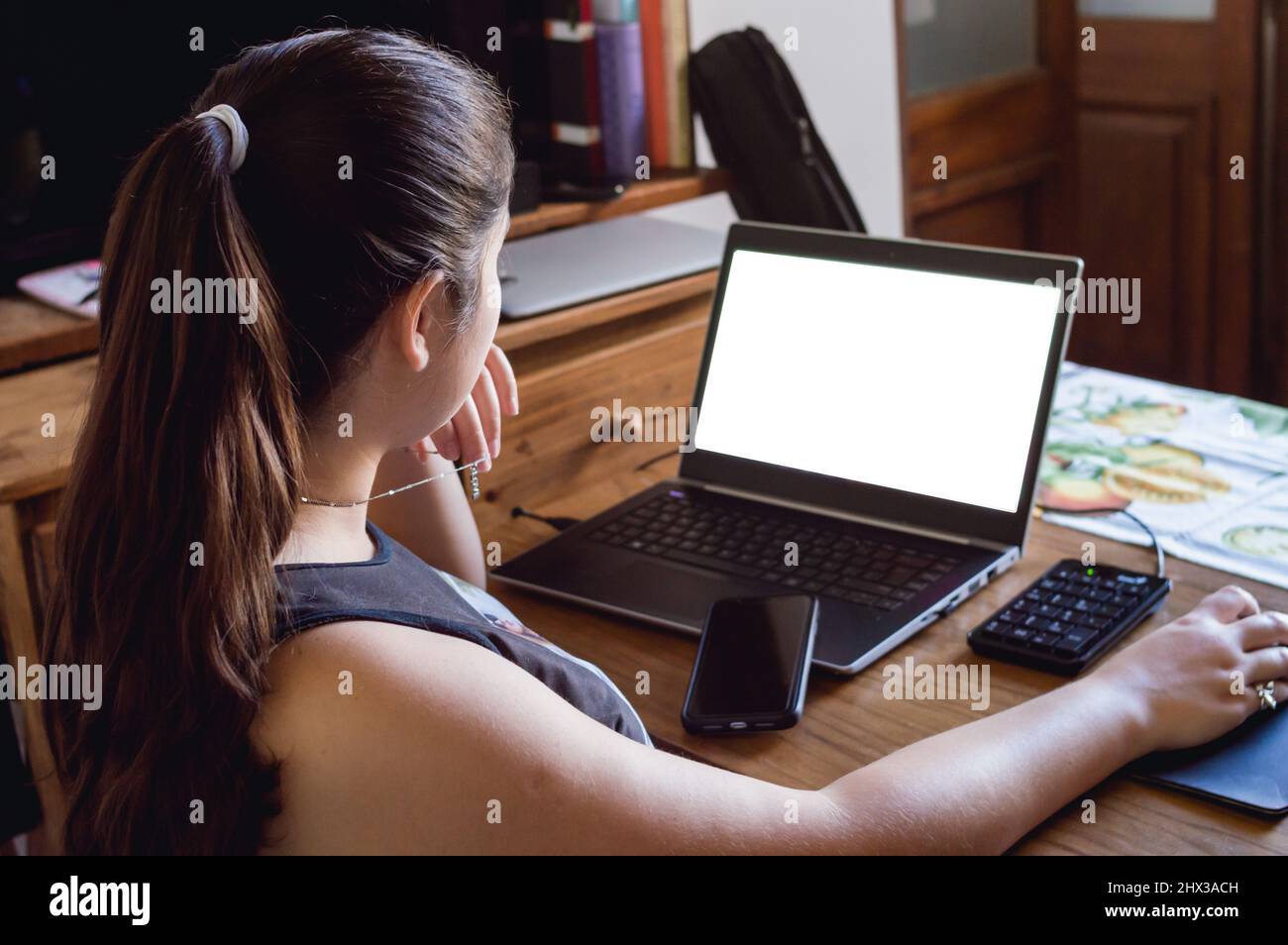 back view of young caucasian woman at home looking at her laptop screen with one hand on the mouse and a cell phone on the keyboard. Stock Photo