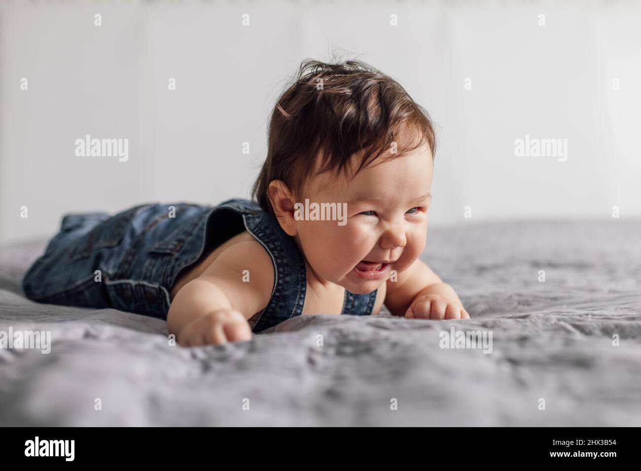 Portrait of pretty baby wearing denim romper lying on gray blanket at home. Crying infant child crawling on bed in bedroom, white background Stock Photo