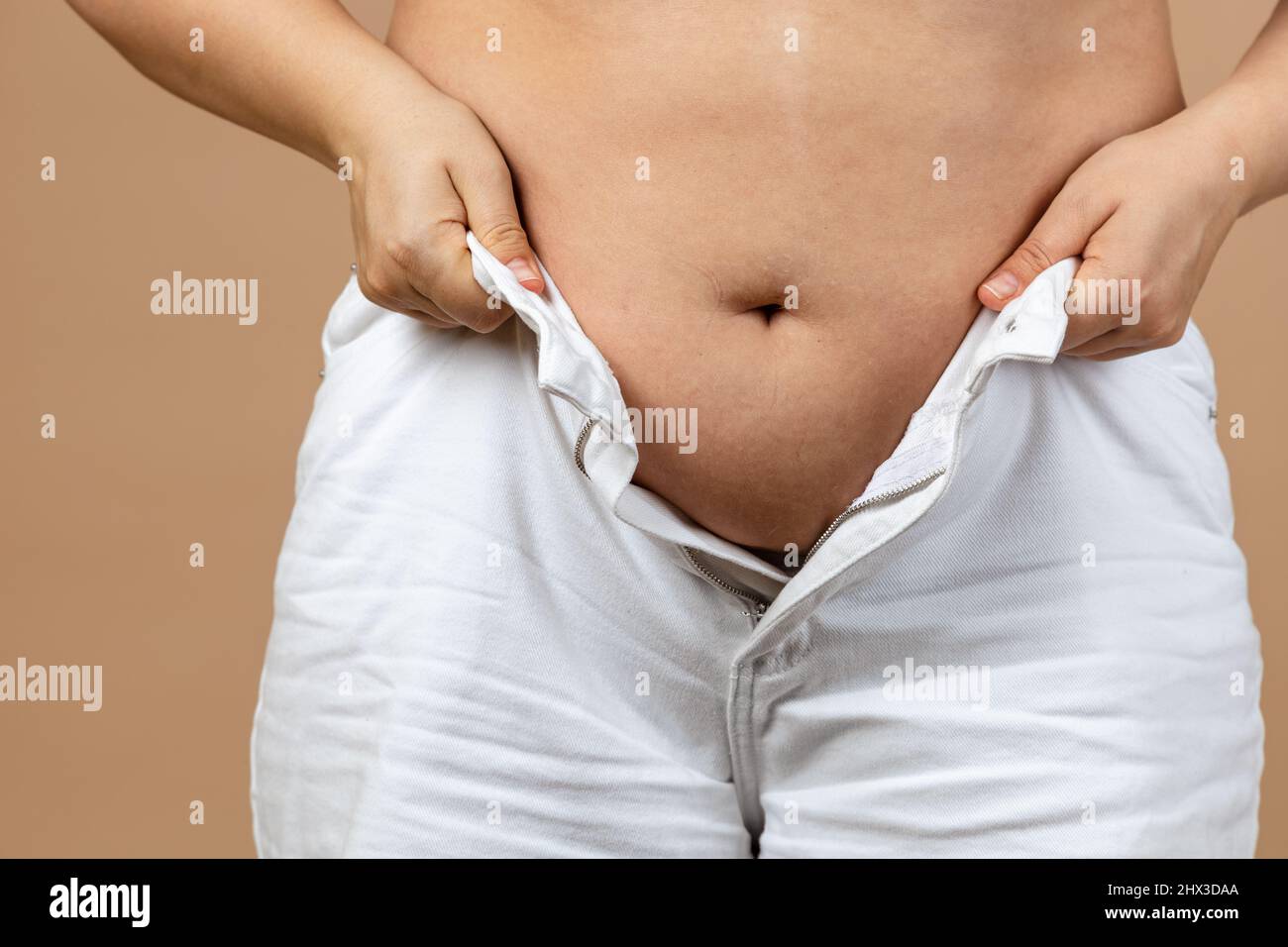Closeup of lady with thick plumpy abdomen trying to zip up white shirts on beige background. Visceral fat. Body positive. Sudden weight gain. Tight Stock Photo