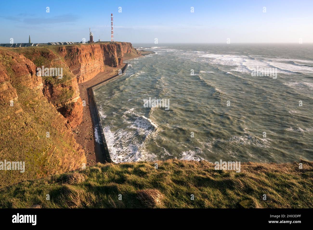 Tall red dramatic cliffs of Heligoland island with rough sea and a lighthouse far in the back. Sunny windy winter day in Helgoland in the North sea Stock Photo