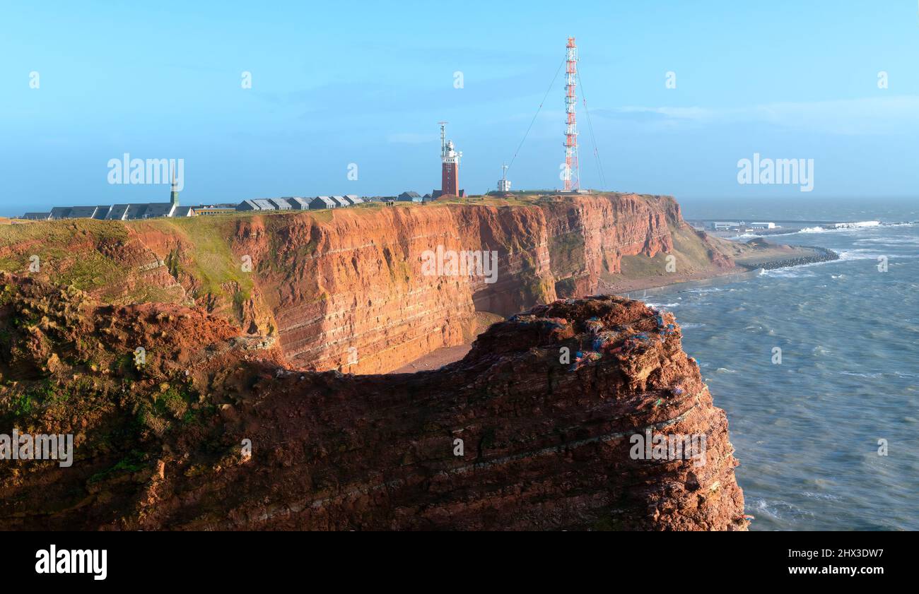 Tall red dramatic cliffs of Heligoland island with rough sea and a lighthouse far in the back. Sunny windy winter day in Helgoland in the North sea Stock Photo
