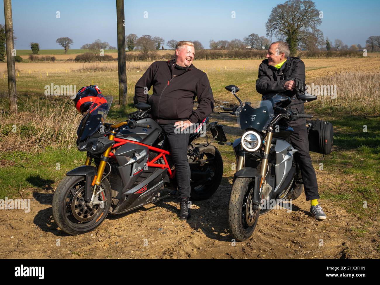 Two middle aged men motorcyclists sit on Energica EV electric motorcycles as they chat during a rest from riding. Stock Photo