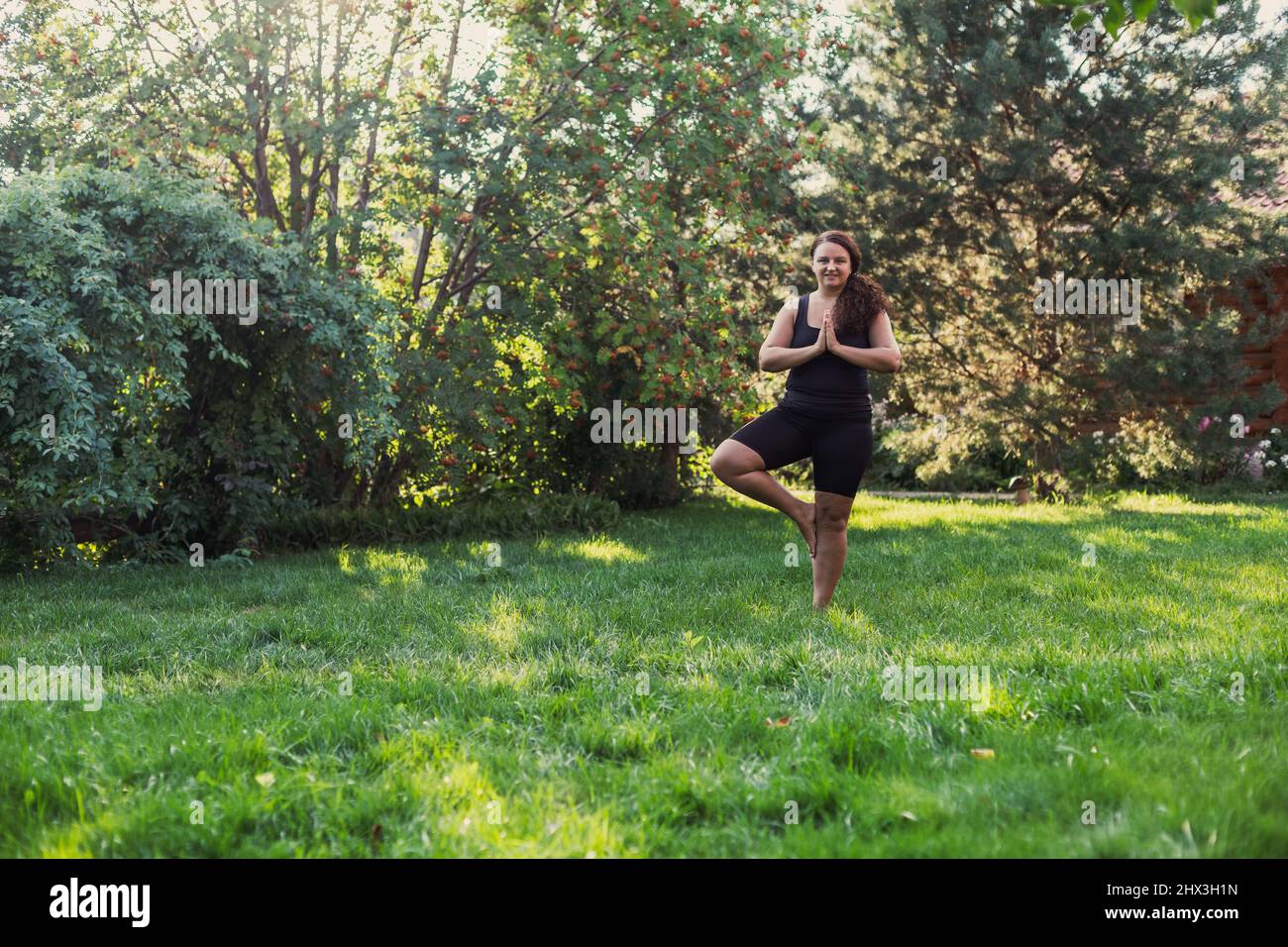 Female with extra weight standing on one foot in yoga position enjoying the moment on green grass on backyard of cottage with wooden house and trees Stock Photo