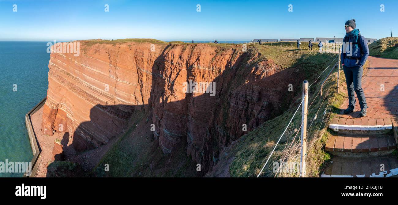 Woman standing on the edge of tall red dramatic cliffs of Heligoland island with rough sea. Sunny windy winter day in Helgoland in the North sea Stock Photo