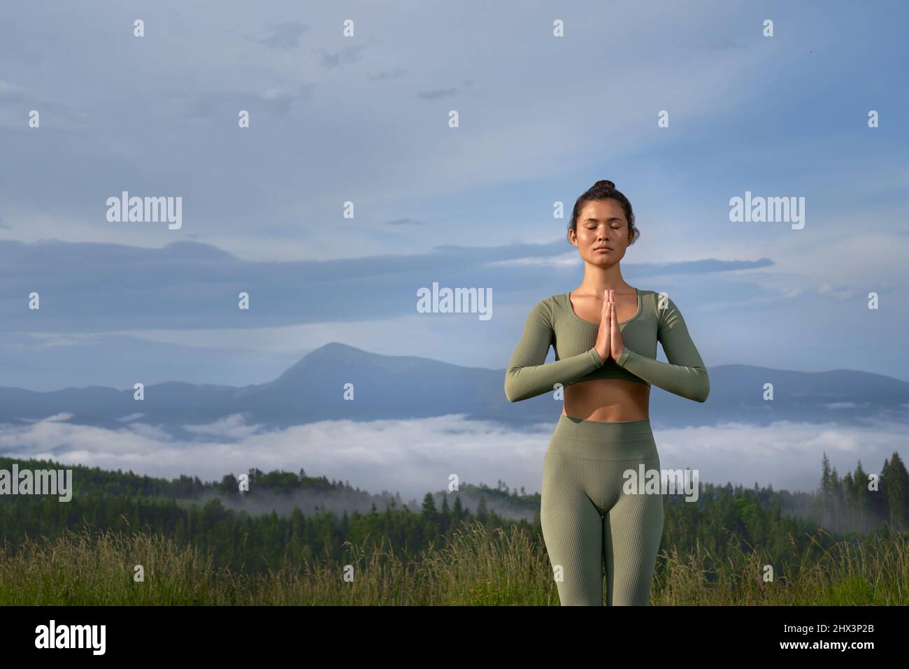 Beautiful young woman meditating with closed eyes with background of green mountains. Peaceful lady standing with namaste gesture and enjoying harmony on nature. Stock Photo
