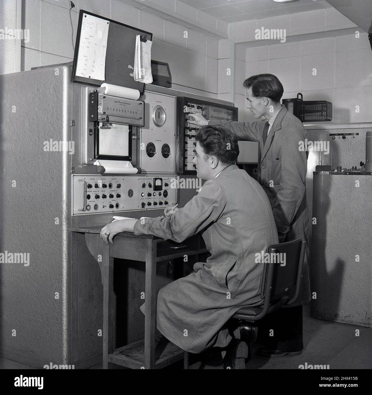 1950s, historical, two male technical staff in long work coats at the control panel of a Spectrometer at the steelmaking factory at Abbey Works, Port Talbot, Wales, UK, one seated at a small desk, the other standing. Both are studying the graphic data being displayed on the machine's print-out. Due to the numeous metals used, steel making requires accurate and precise analyse and a production and monitoring spectrometer as seen here - made by Applied Research Laboratories of Glendale, CA -  was used in the industry at this time to ensure a constant output. Stock Photo