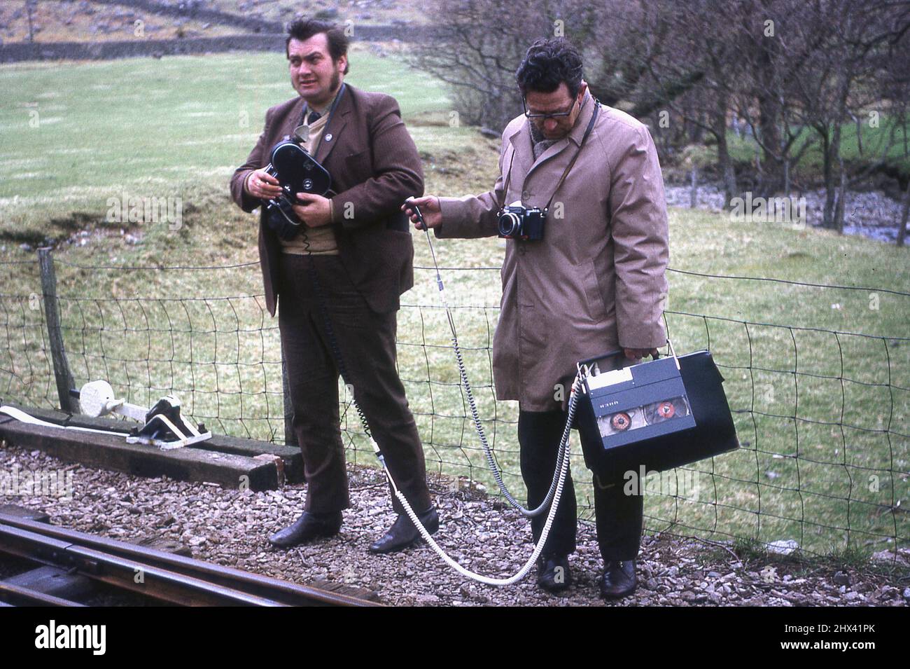 1971, May, a film cameraman and a sound or audio recordist, on location, standing beside a railway track, England, UK. The sound recordist is holding a portable reel to reel sound recording machine and microphone to record the surrounding sounds. Stock Photo