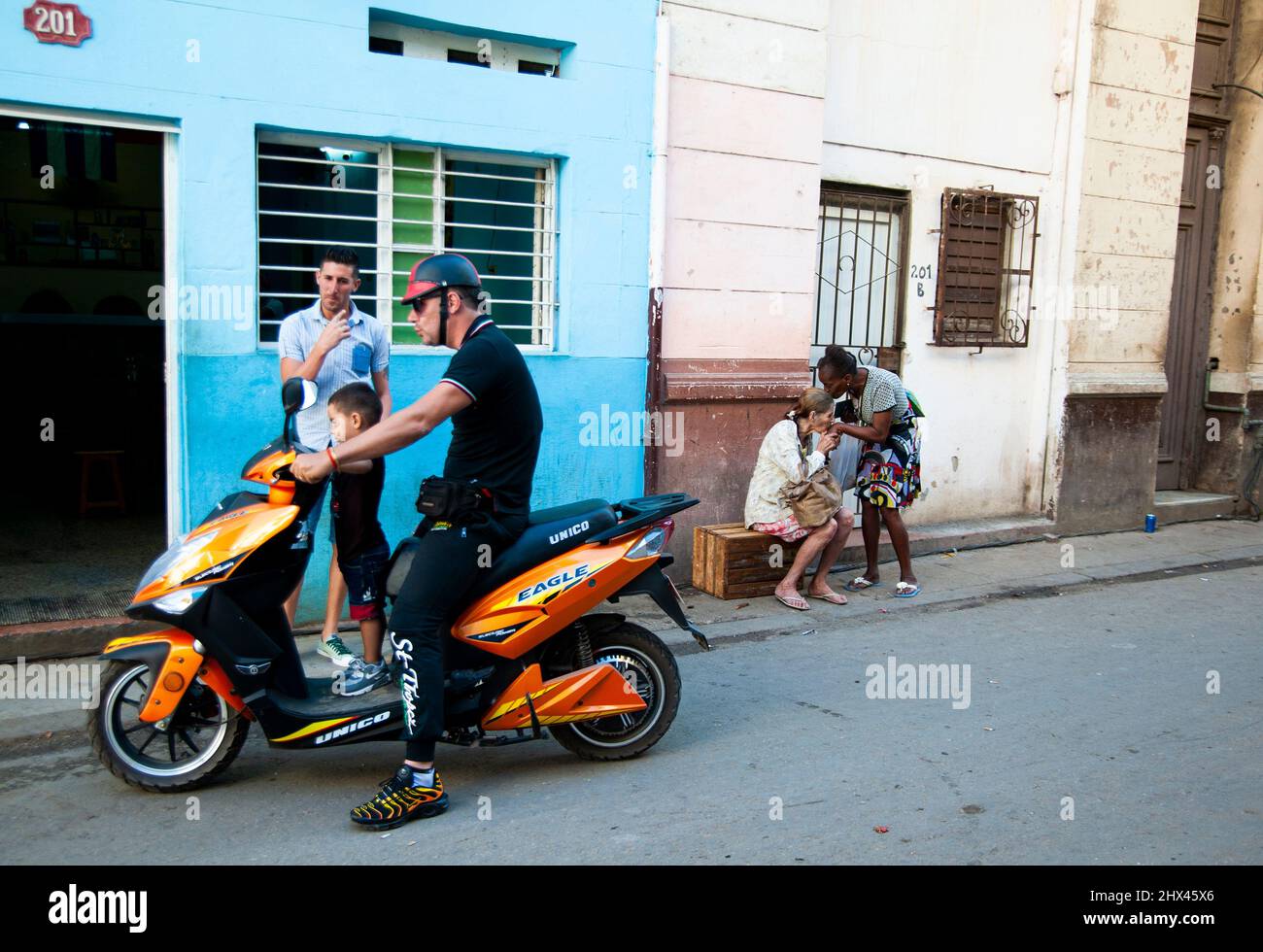 Two women embrace each other with a kiss while a man with his son play on a motorcycle in Havana, Cuba. Stock Photo