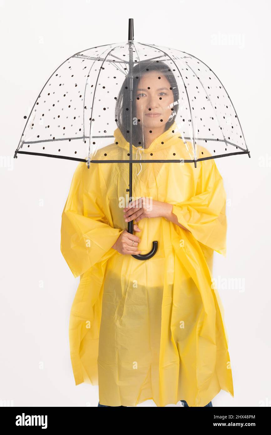 Studio shot of beautiful Asian woman with long black hairs wearing yellow raincoat and hiding under transparent with black dots umbrella with copy spa Stock Photo