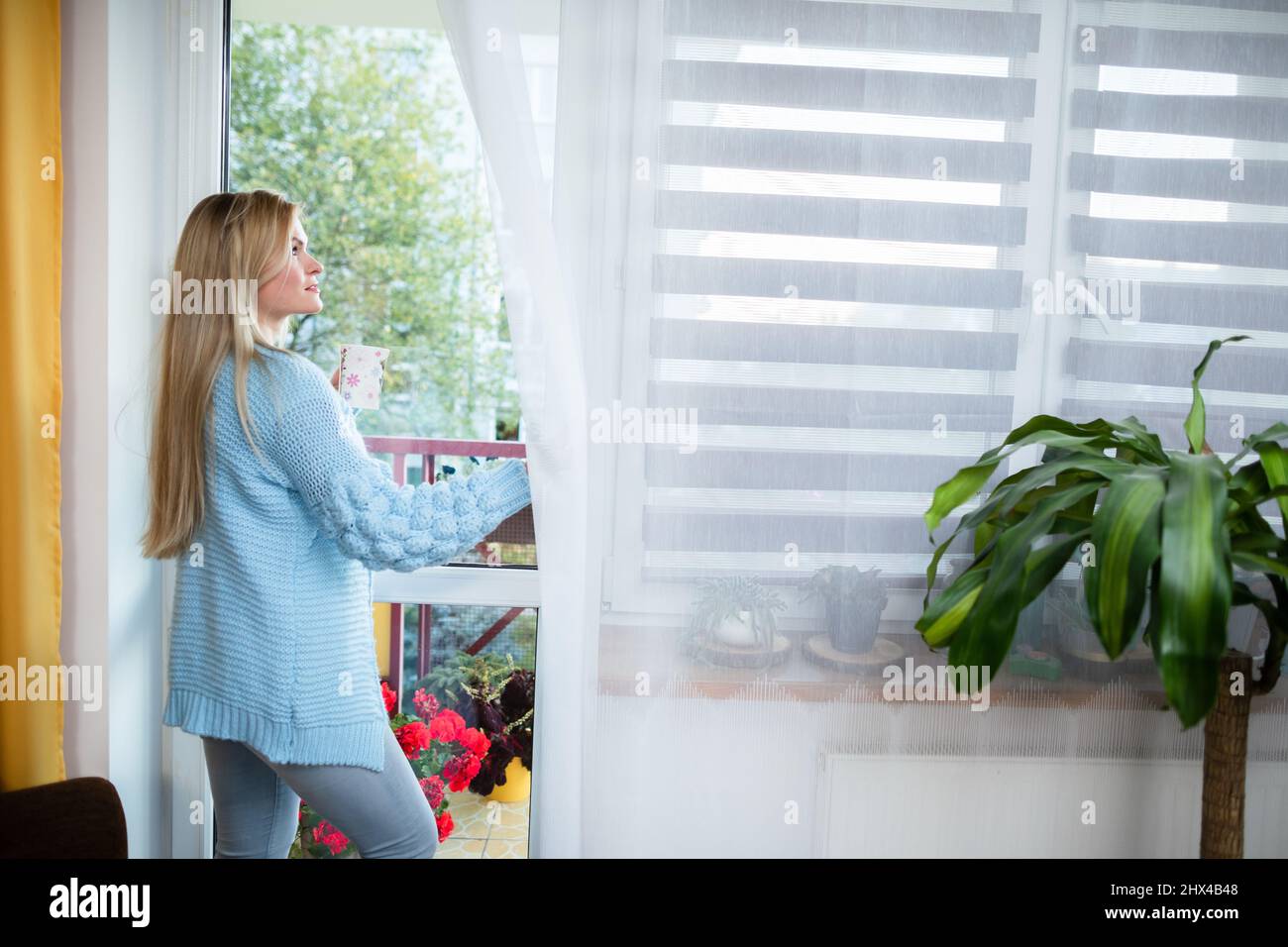 The girl looks outside through the window and her hands hold a cup. Stock Photo