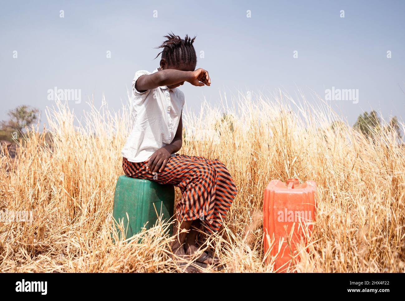 Exhausted little girl sitting on a plastic water canister in a dry field in the African bush suffering from scorching heat and her hard work; Child la Stock Photo