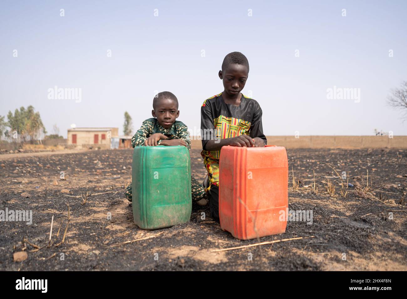Two sad African boys kneeling in a burnt field behind their large water canisters, staring desperately at the ground in front of them; Water shortage Stock Photo
