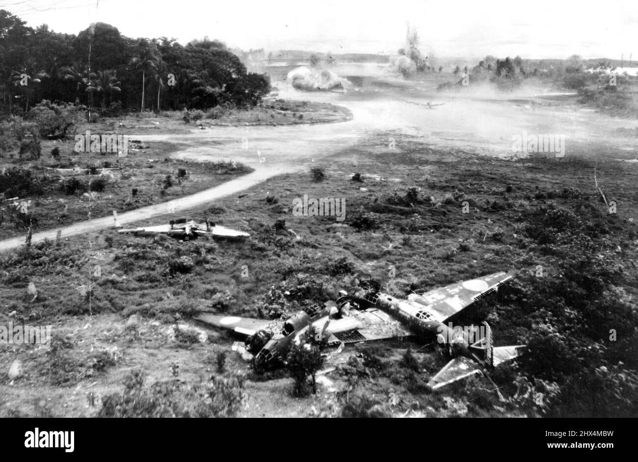 Fifth Japanese Planes Destroyed -- In a swift, devastating strike by U.S. Fifth Air Force bombers this last effective Nip air base in New Guinea recently was badly damaged. The location is Jefman airdrome, Sorong Island, where 50 Japanese planes were destroyed both on the ground and in the air. In this photo two Nip planes are seen destroyed on the ground while a third rises to a sudden, fiery end in the air. When the U.S. bombers had finished their work, not a serviceable Jap plane remained on the ground. Note the delayed bomb being parachuted to the ground right on the tail of the Jap bombe Stock Photo