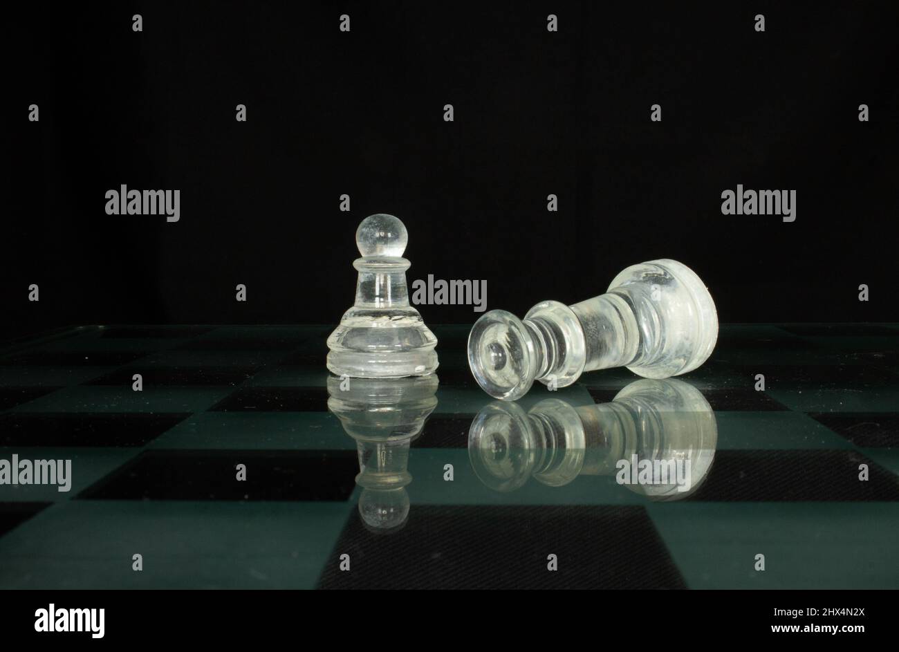 clear chess piece pawn and fallen queen isolated on a chequered chess board with a black background Stock Photo