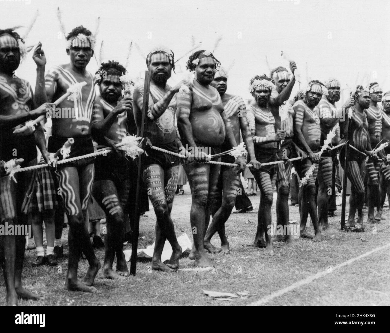 Native skin colouring - maggie brown to Kurir black designer our bodies - mainly yellow ochre - some red coin cloths - deep red, nearly black. White rope held up by black metre pickets are holding bones decorated with with tosses. At Whyalla aborigines from a mission station "dressed" for the Queen's visit. April 12, 1954. Stock Photo