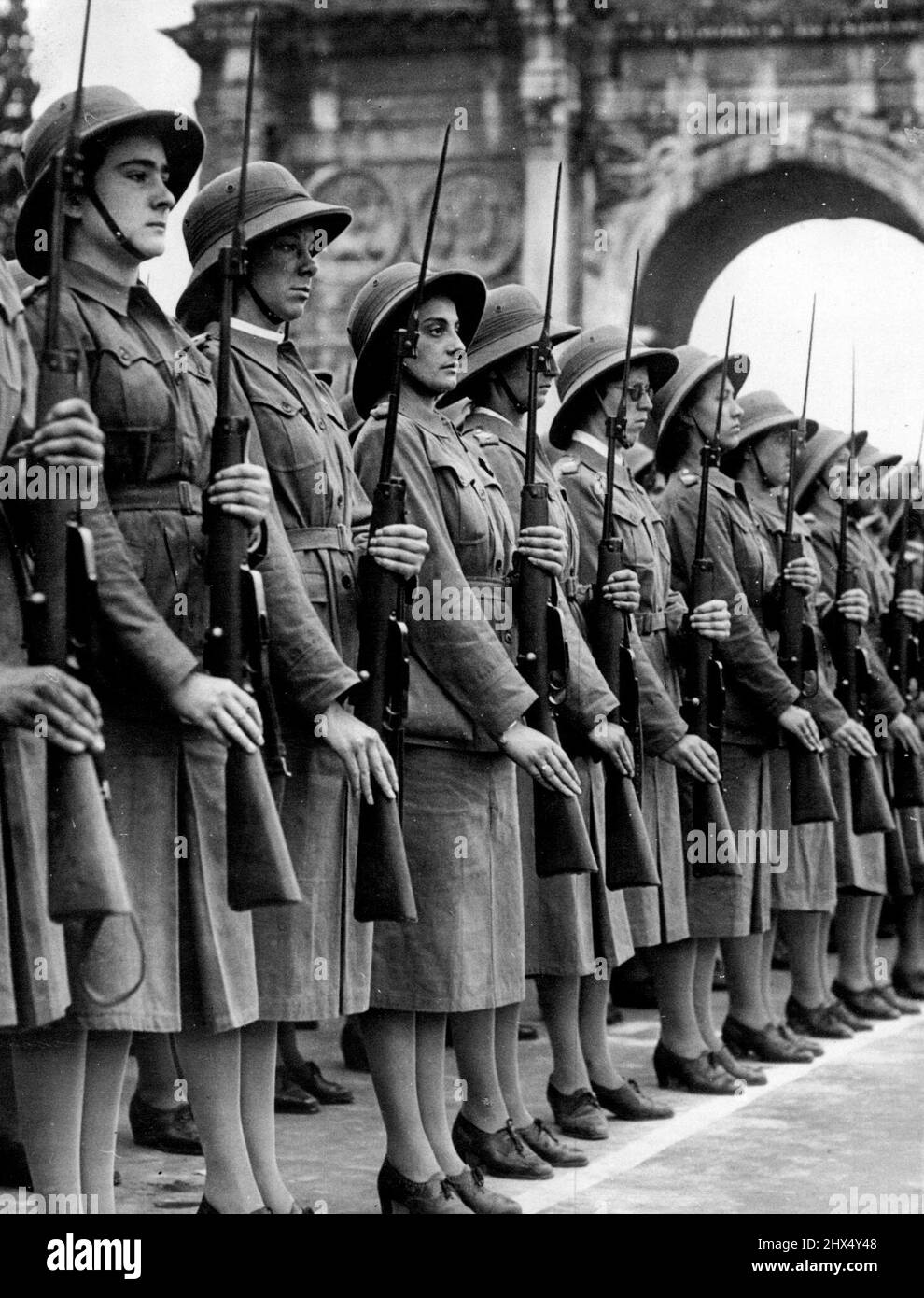 Khaki-dressed girls in sun helmets and carrying rifles with fixed bayonets stand at attention after marching past Signor Mussolini in Rome, where 70,000 Fascist women and girls took part in a parade for their leader. June 10, 1939. (Photo by The Associated Press of Great Britain Ltd.) Stock Photo