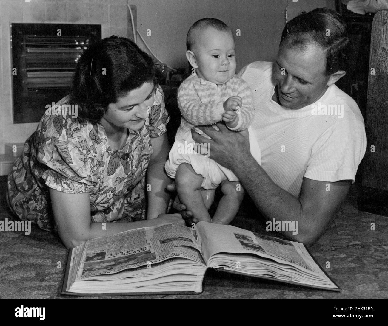 Bob and Mrs. Scott look through the scrapbook which they kept together of his deeds in Australia, while Bruce jigs happily. Bruce is just starting to climb up onto his feet, but Bob always puts him down. 'I want his legs to be good and straight for goal-kicking,' he explains. July 13, 1951. Stock Photo