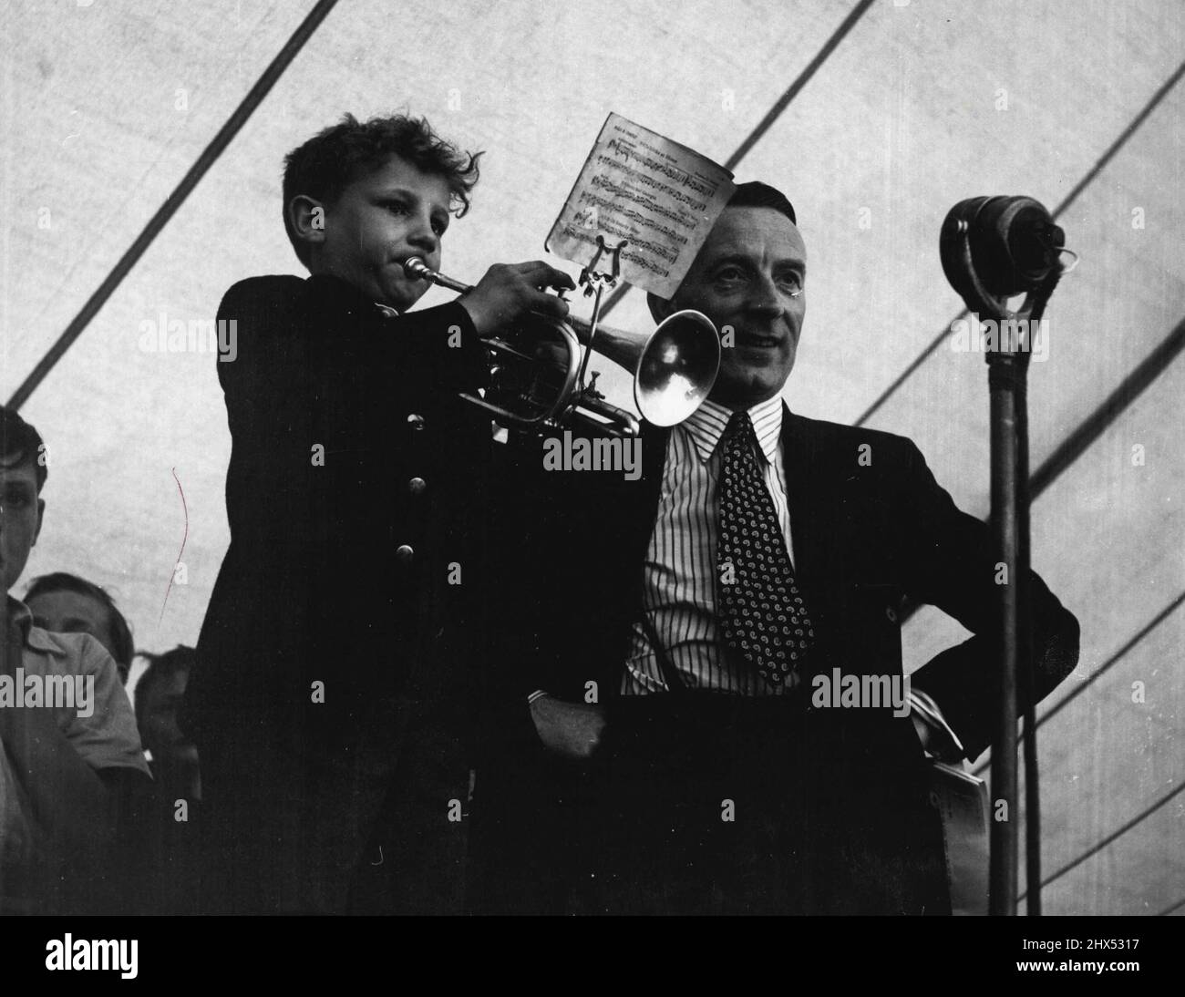 Pickles Party -- 'Ee, that's champion.' Hands on hips, Wilfred listens to the youngest trumpeter in the Church band. He's Arthur Slater, aged nine. ('Are the Coortin'?' asks Pickles.) Arthur plays the Londonderry Air on his cornet. Something goes wrong after a couple of bars. 'He valve's sticking,' says Arthur, to Wilfred and the listeners. 'Never mind,' Wilf reassures him. 'We don't mind a valve sticking here and there! ***** a go!' July 22, 1948. (Photo by Pictorial Press). Stock Photo