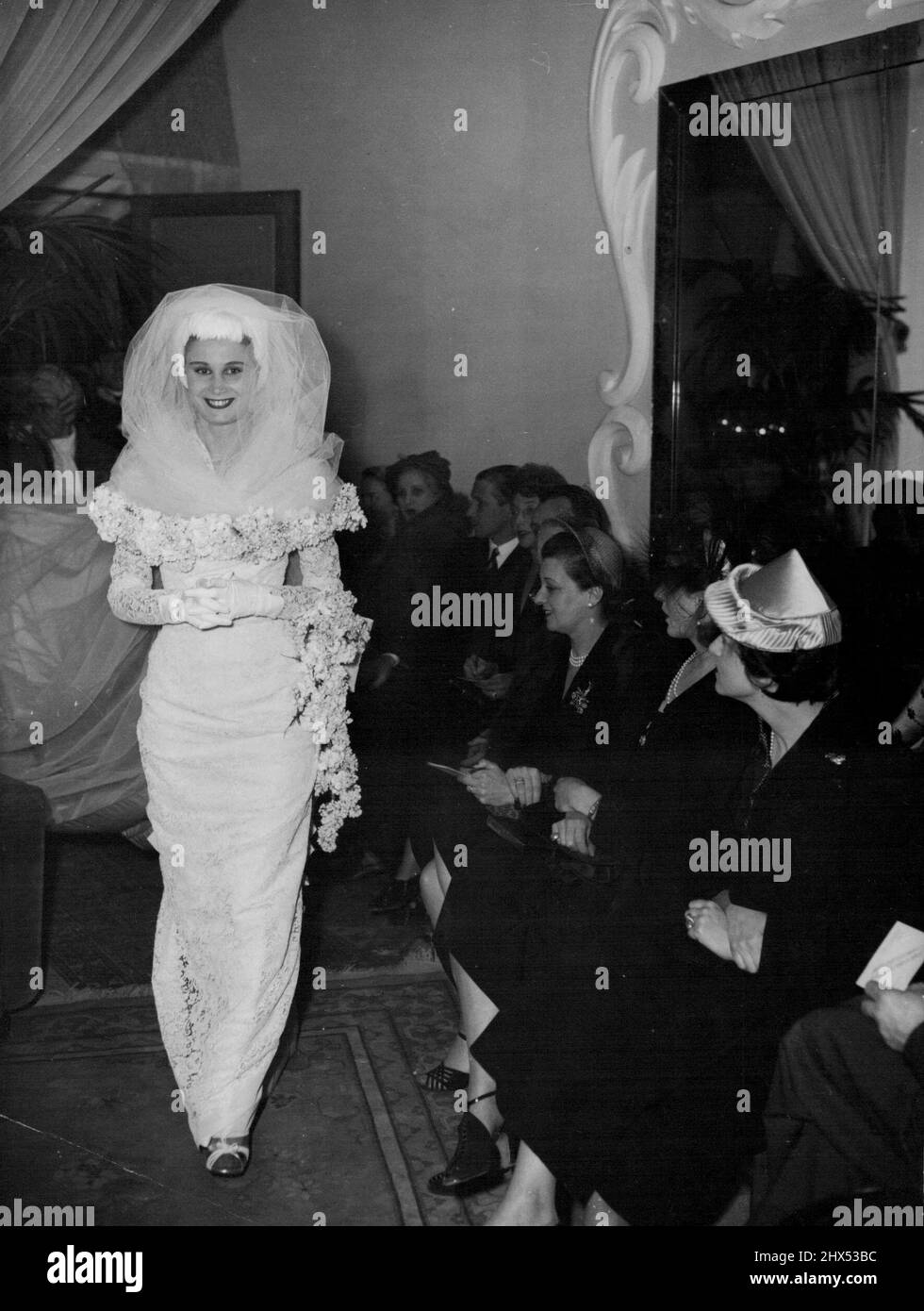 Dress Designer's Story -- This wedding gown opened Schubert's Recent show. It is in heavy Venetian lace, bodice and sleeves Decorated with flowers. A white feather cap and a veil top the outfit. The train was about 13 yards long. Eleven years age Emilio Schuberth started designing hats in Rome. After a year, he opened a fashion house, and within a few months was commissioned to make the wedding dress for H.R.H. Maria of Savoy, Daughter of King Victoria Emanuele III. Since then, he has never looked back, and now his creations take a leading place in the fashions of the World. July 6, 1949. Stock Photo