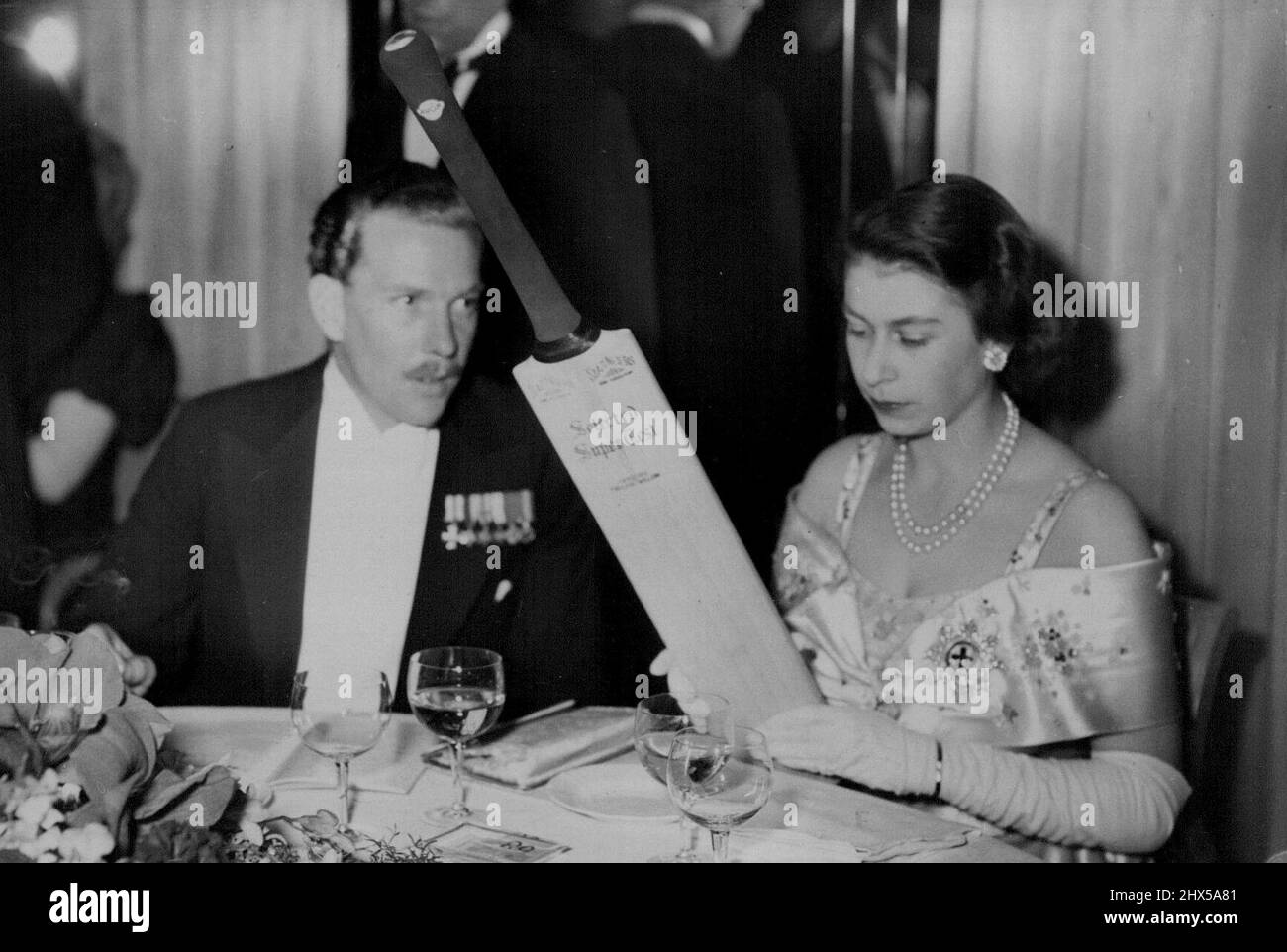The Princess At The Ball - Princess Elizabeth with Major David Butters interested in a cricket bat which was later auctioned on behalf of the Association's funds. Among the guests 'received' by the Duke of Edinburgh at a ball at the Dorchester Hotel in aid of the National Playing Fields Association, of which he is President, was Princess Elizabeth. The Princess, wore the emblem of the Order of the Garter on an off-the-shoulder gown in off-white, embroidered with raised blue crystal beads. April 21, 1949. Stock Photo