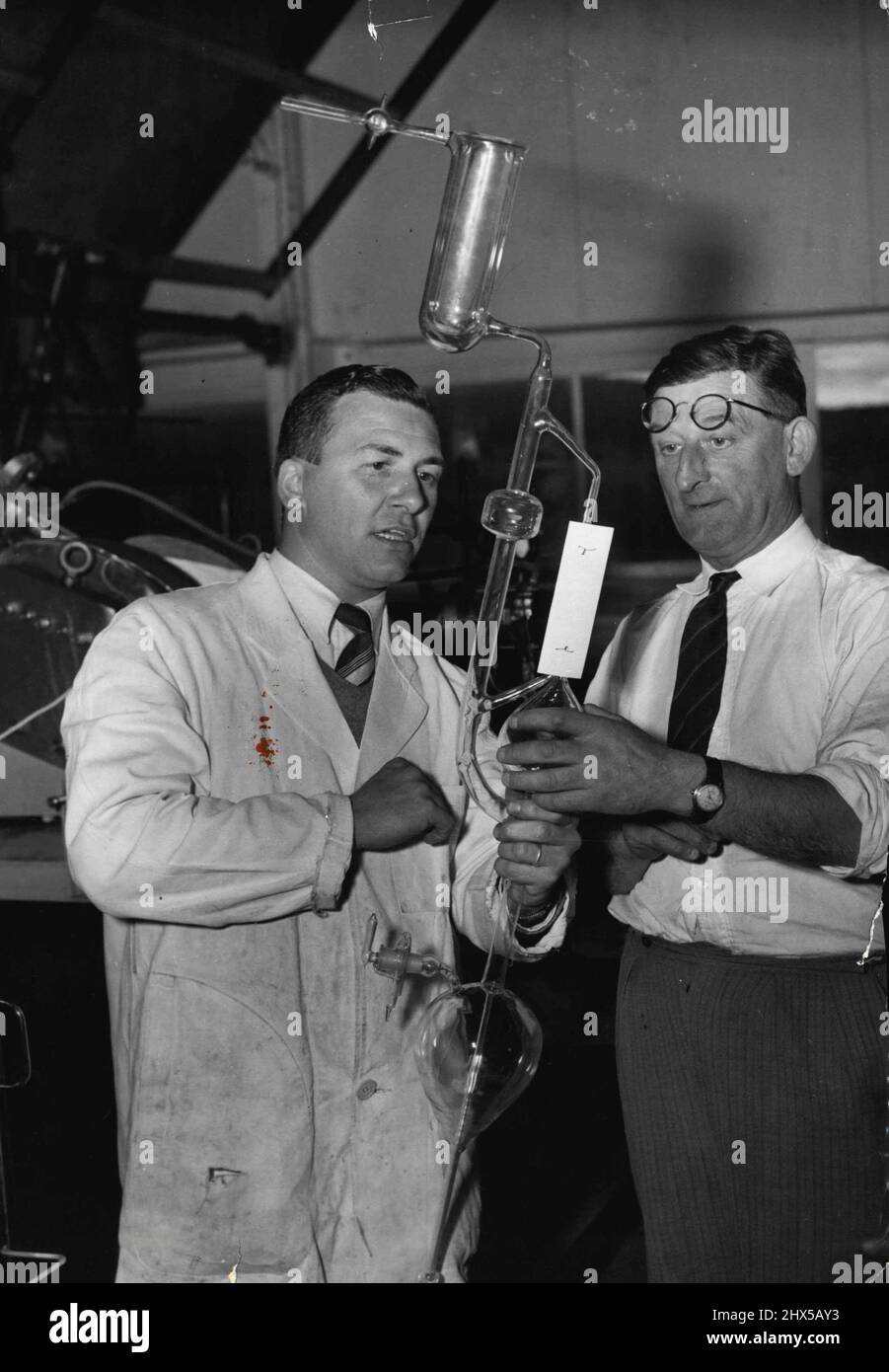 The skill of master glassblower Wilhelmus Tys (left) produced the complicated apparatus he shows her to chief technical officer Mick Cormick of the school of Physical Science, Canberra. November 17, 1954. Stock Photo
