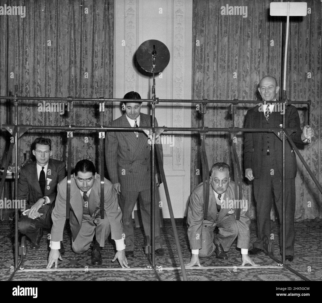 ***** Gate For Trackmen William Moore (front, left) of Louisville, Kentucky, vice-president of the Kentucky AAU, and Wood Hardin, of ***** Albany, Indiana, Kentucky AAU' president, kneel in starting position as they demonstrate a new-type starting gate for runners at the annual convention of the Amateur Athletic Union at the Hotel Astor, New York, Dec. 3. The gate, similar to the starting gates used in horse racing, is designed to eliminate flase starts. Runners lean against straps which swing upward when start is given by a tripping mechanism which also fires a gun at the top as a timer's sig Stock Photo
