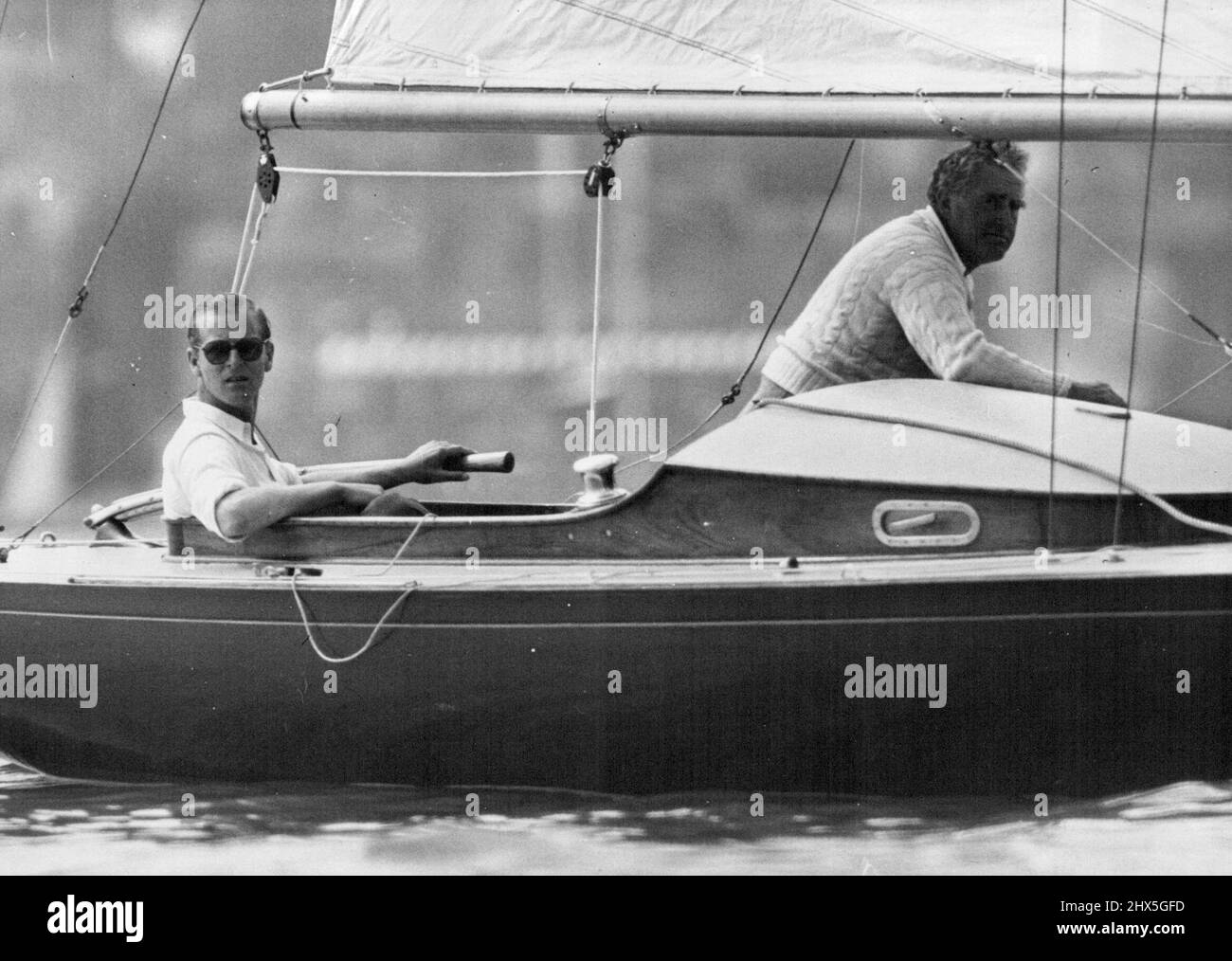 The Duke Becalmed In Cowes Race -- The Duke of Edinburgh (left) at the tiller and Mr. Uffa Fox (designer of the Coweslip) photograph had becalmed during the race at Cowes today. The Duke of Edinburgh taking part in the Cowes Regatta, today helped to crew his yacht Bluebottle, which he shares with the Queen in the Dragon Class race. August 6, 1952. (Photo by Fox Photos). Stock Photo