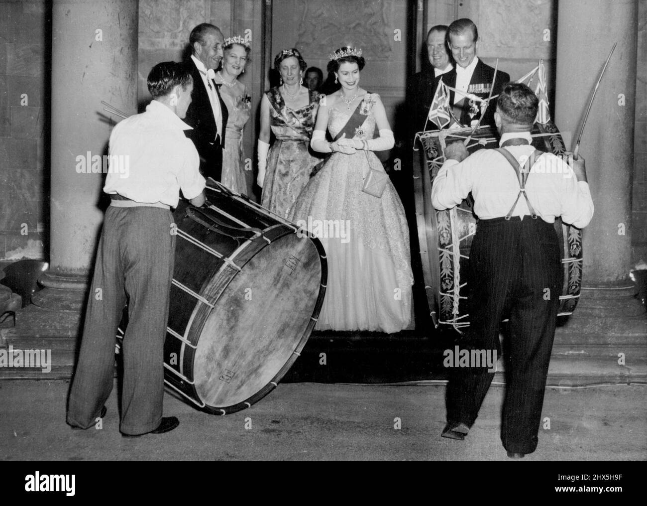 Drums Beat For The Queen. The Queen and the Duke of Edinburgh smile happily as they watch Irish drummers beating outside drums with canes outside Government House. Earlier the Royal couple had arrived at Hillsborough for their three-day visit to Northern Ireland. Standing beside the Queen are, left to rights Viscount Brookeborough, Prime Minister of Northern Ireland, Lady Wakehurst, wife of the Governor, and Lady Brookeborough. July 2, 1953. (Photo by Planet News Ltd.). Stock Photo