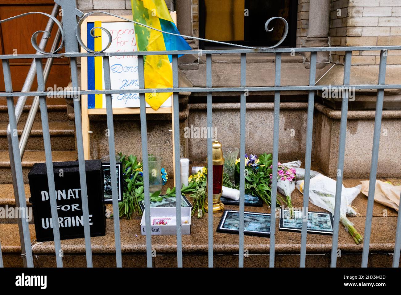 New York, NY, USA. 9th Mar, 2022. A banner asking people to pray for Ukraine, a box for donations, flowers, photos, a votive candle, and a Ukrainian flag and memorabilia sit in the rain outside the Holy Trinity Ukrainian Orthodox Cathedral in New York City's Little Italy. Credit: Ed Lefkowicz/Alamy Live News Stock Photo