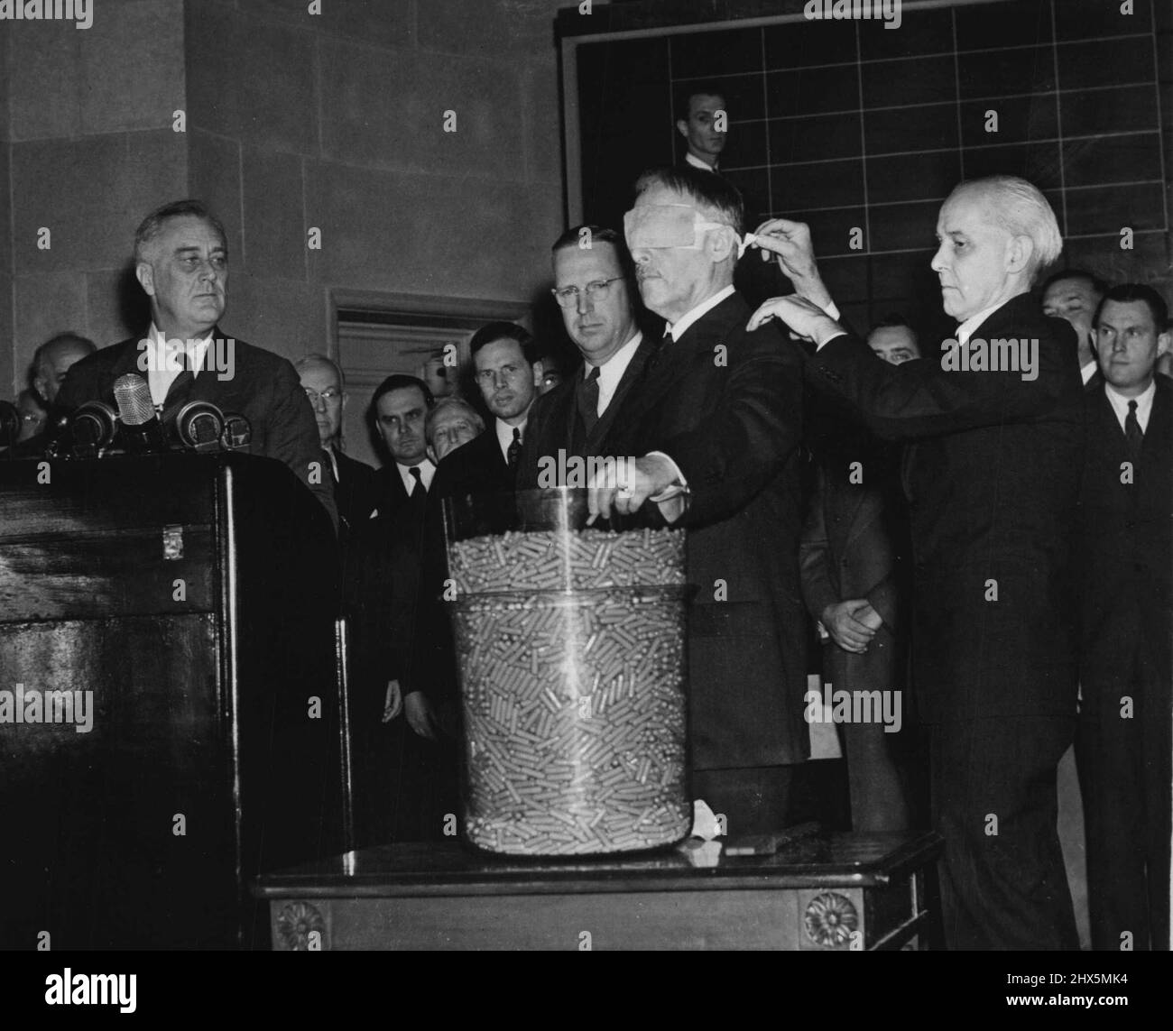 Stimson Draws First Draft Capsule -- The camera records an epochal moment in the history of the United States as secretary of War Henry Stimson, Blindfolded, draws the first number in the first peace time draft lottery in Washington on Oct. 29th. President Roosevelt is looking on at left; and Colonel Charles R. Morris, retired, who Blindfolded officials in the World War draft, is at secretary Stimson's shoulder. The number drawn by Stimson was 158. October 29, 1940. (Photo by ACME). Stock Photo