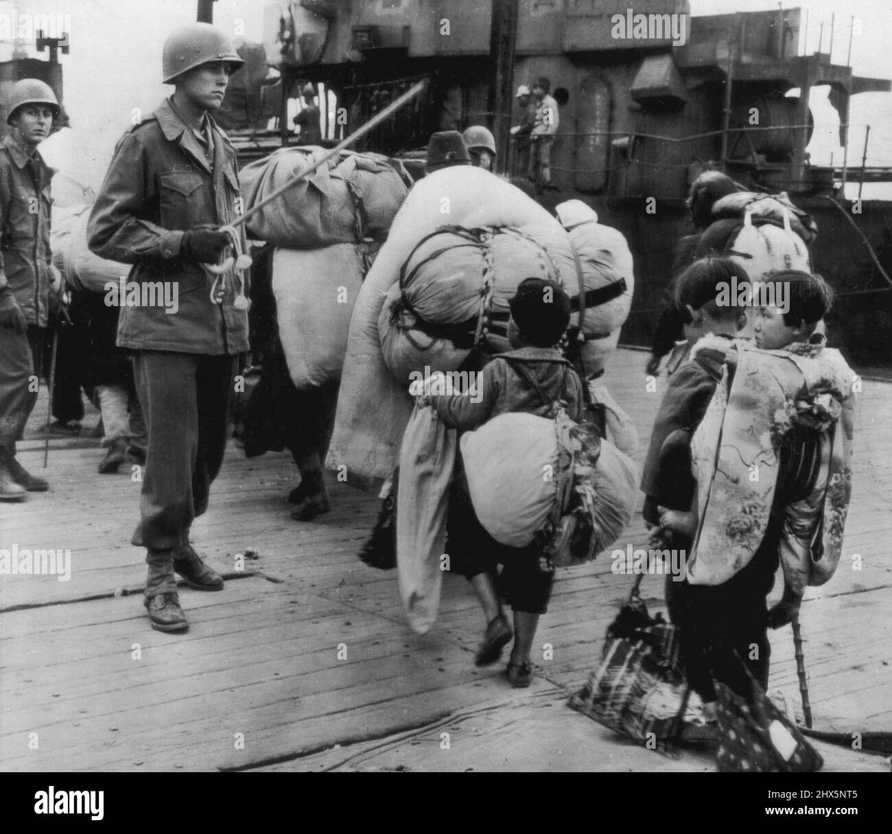 Japs Go Home with Heavy Burdens - A Japanese child Laden with Bundles and carrying her Tiny Brother on her back trods across a pier in Fusan, Korean Port, with other Burdened Japanese being repatriated to their homeland. December 11, 1945. Stock Photo
