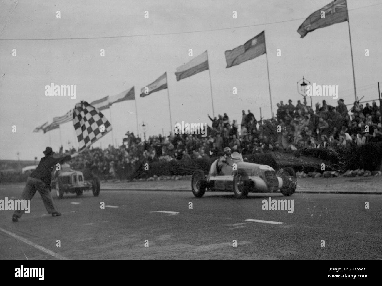 ***** International Road Race -- Bob Gerard, the winner, being flagged as he finishes. The car on the right, which appears to be leading. ***** laps to complete, it is the ***** by Prince Birabongse. Bob Gerard, of Leicester in his E.R.A. to-day ***** the Jersey International 176-mile ***** race of 55 laps at St. Helier. He ***** two laps ahead of G. Abecassis, ***** was second in a Megerati. Reg. ***** year's winner, was third and Prince Birabongse (B. Bira), was fourth, they both drove Masers Stock Photo