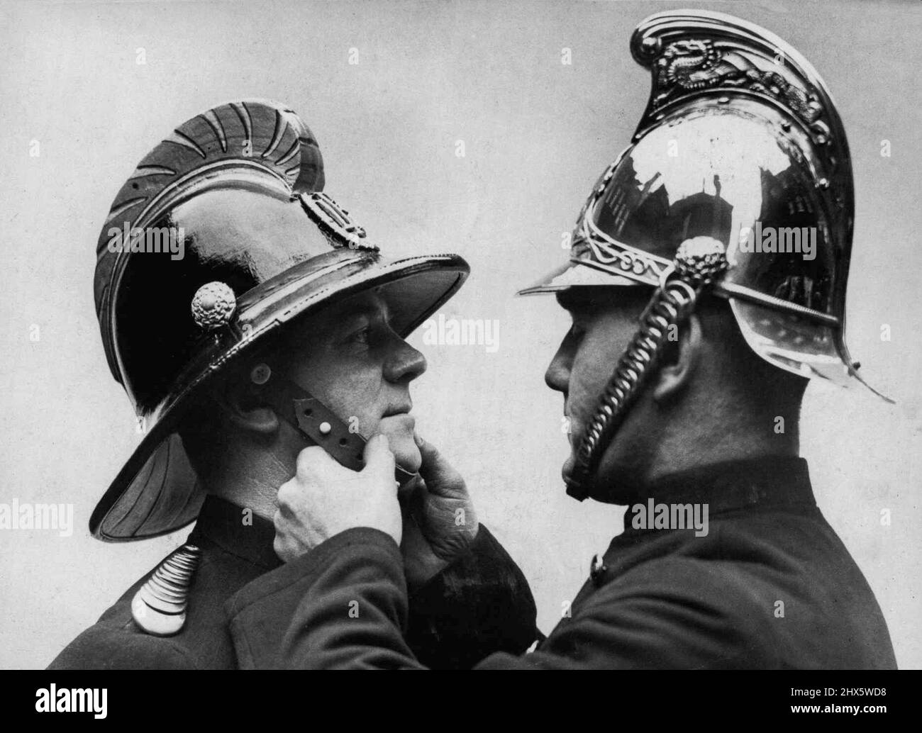 New Helmet for Firemen. A fireman wearing the old type helmet adjusts the strap of a new model. The seining brass helmet of the London fire Brigade, introduced over sevent years ago by Sir Eyre Massey Shaw, the first chief officer of the Brigade, replaced the old-time leather helmet is, in its turn, being superceded. The new helmet of cork and rubber is glossy black with gilt comb and rosettes, silver in the case of principal officers. Although the brass helmet gave a high measure of protection, it failed in these modern times through its high conductivity of electricity. November 21, 1936. Stock Photo