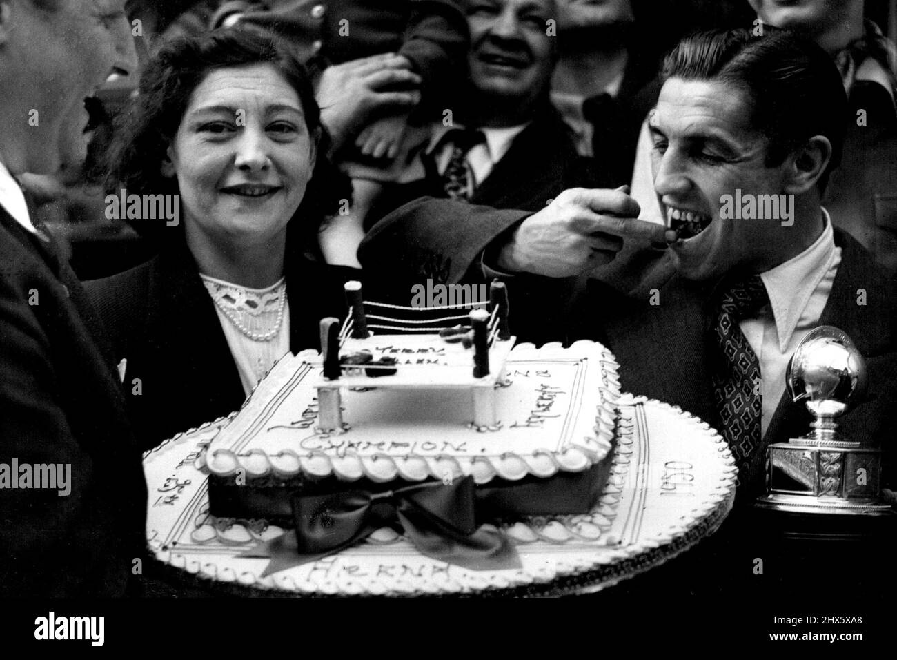 New Champion Takes The Cake - Terry Allen, new world flyweight champion, can't wait to take a taste of this huge cake, appropriately ornamented with a boxing ring, presented to him to-day (Monday) in Chapel Street market, Islington, North London - where he was once a barrow boy. At left is Allen's wife.The market was crowded for the presentation of the cake, given by a local baker. May 1, 1950. (Photo by Reuterphoto). Stock Photo