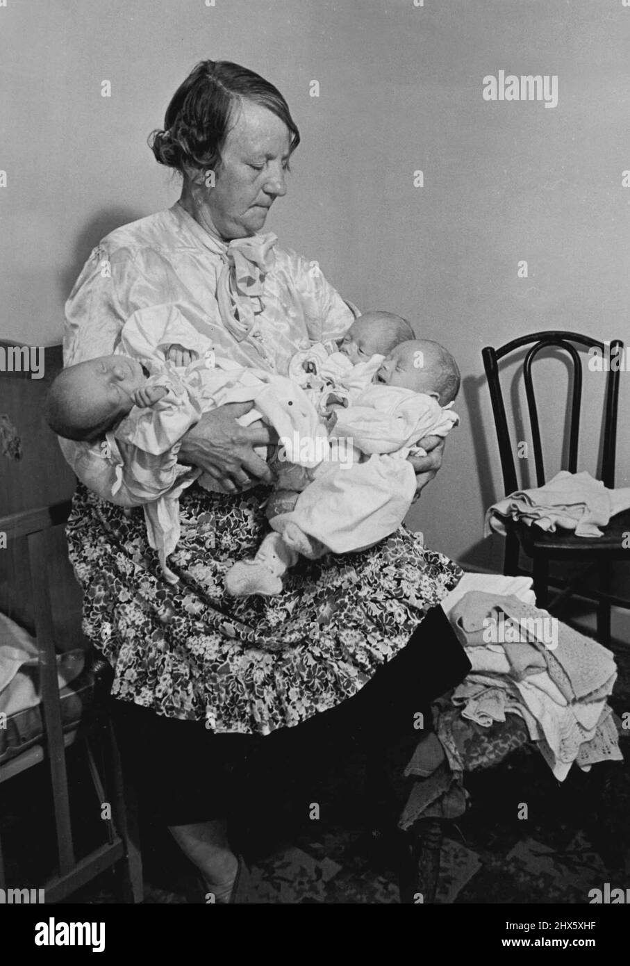 Granny Is Home Help -- Granny holds the triplets while the mother, Mrs. Joyce Palmer, sorts out their clothes.There's nothing unusual in granny helping to look after the babies, especially when they are triplets, but this granny is paid 1s.10d. an hour by Essex County Council to do it. Granny Mrs. Edith Alice Adams, is the mother of the triplets mother, Mrs. Joyce Palmer, 22, of The Lowe, Chigwell, Essex, The triplets all boys, are three weeks old. Their names right to left are Stock Photo