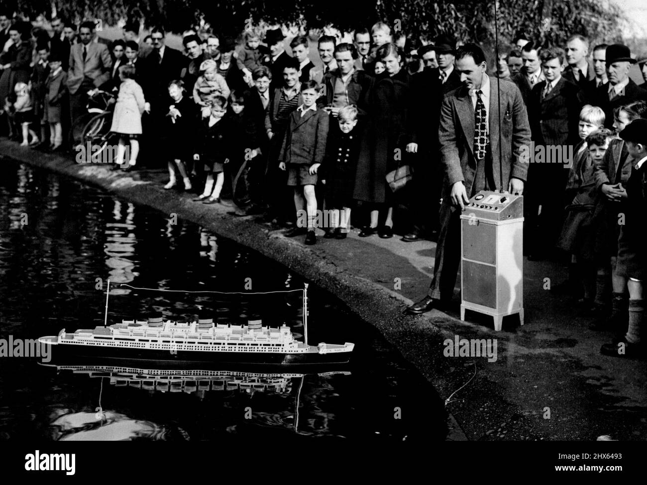 This Queen Mary Has Radio Control -- Ashore the radio controlled Queen Mary attracts a large crowd as Mr. Pyser removes the 'upper deck' at prince of Wales' pond, Blackheath. Model boat enthusiasts and pool side strollers got a surprise the other day when they saw a perfect scale model of the liner 'Queen Mary' ploughing steadily across the 'Ocean' of prince of Wales Pond, Blackheath. The ship turned, went astern and even hove to off shore, with only the radio antennae to give a clus as to how i Stock Photo